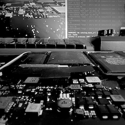 black and white image of a computer circuitboard in the foreground, the background features a screen with some CLI instructions